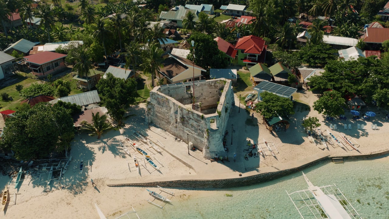 Old stone watchtower ruins beside tropical beach with outrigger boats and village houses Pamilacan Island Bohol, aerial orbit