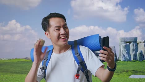 close up of asian male hiker with mountaineering backpack waving his hand and pointing back while having a video call on smartphone while traveling in stonehenge