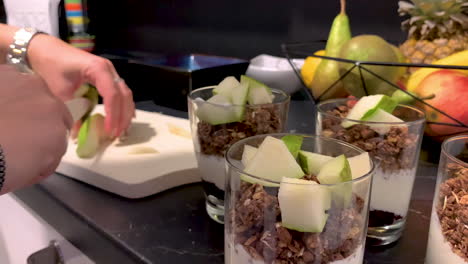 the girl is holding a knife in her hand and cutting a pear on a wooden board, next to her there are glasses of yoghurt and flakes