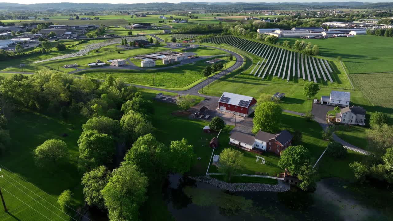 Solar panels farm on green pasture near Wastewater Treatment Plant in American city. Aerial forward wide shot. Production of green renewable energy. Green trees blooming in spring at Sunset.
