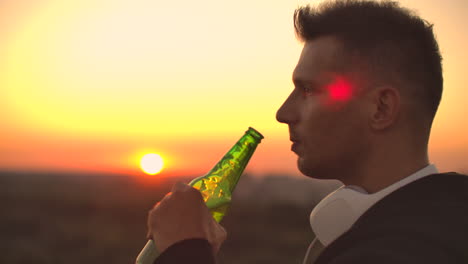 Primer-Plano-De-Un-Hombre-Bebiendo-Cerveza-Al-Atardecer-Parado-En-El-Techo-De-Un-Edificio-Con-El-Telón-De-Fondo-De-Una-Hermosa-Ciudad-Nocturna.