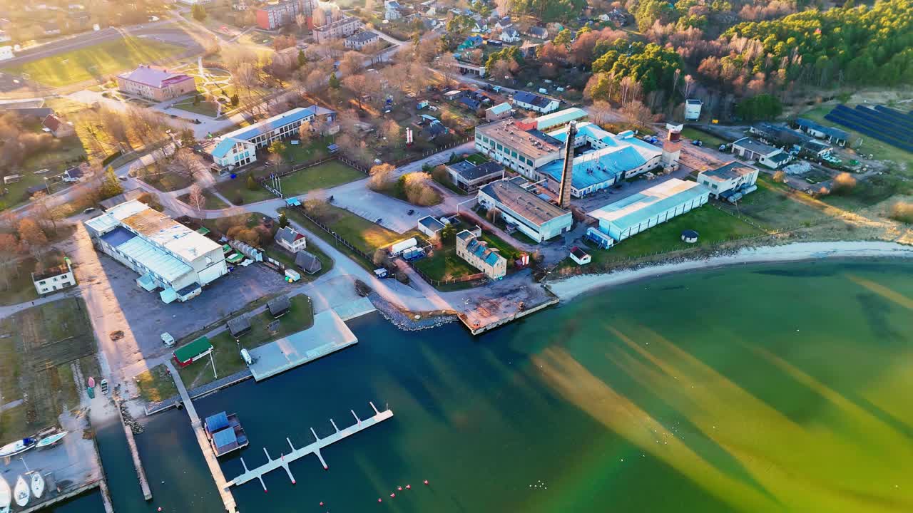 Coastal industrial complex with docks and green waterfront under sunset shadows – aerial view