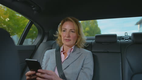 mature businesswoman sitting in back seat of taxi or car checking messages on mobile phone