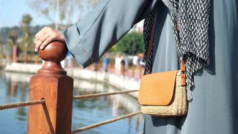 a young woman in a hijab and stylish outfit stands on a walkway near a body of water.