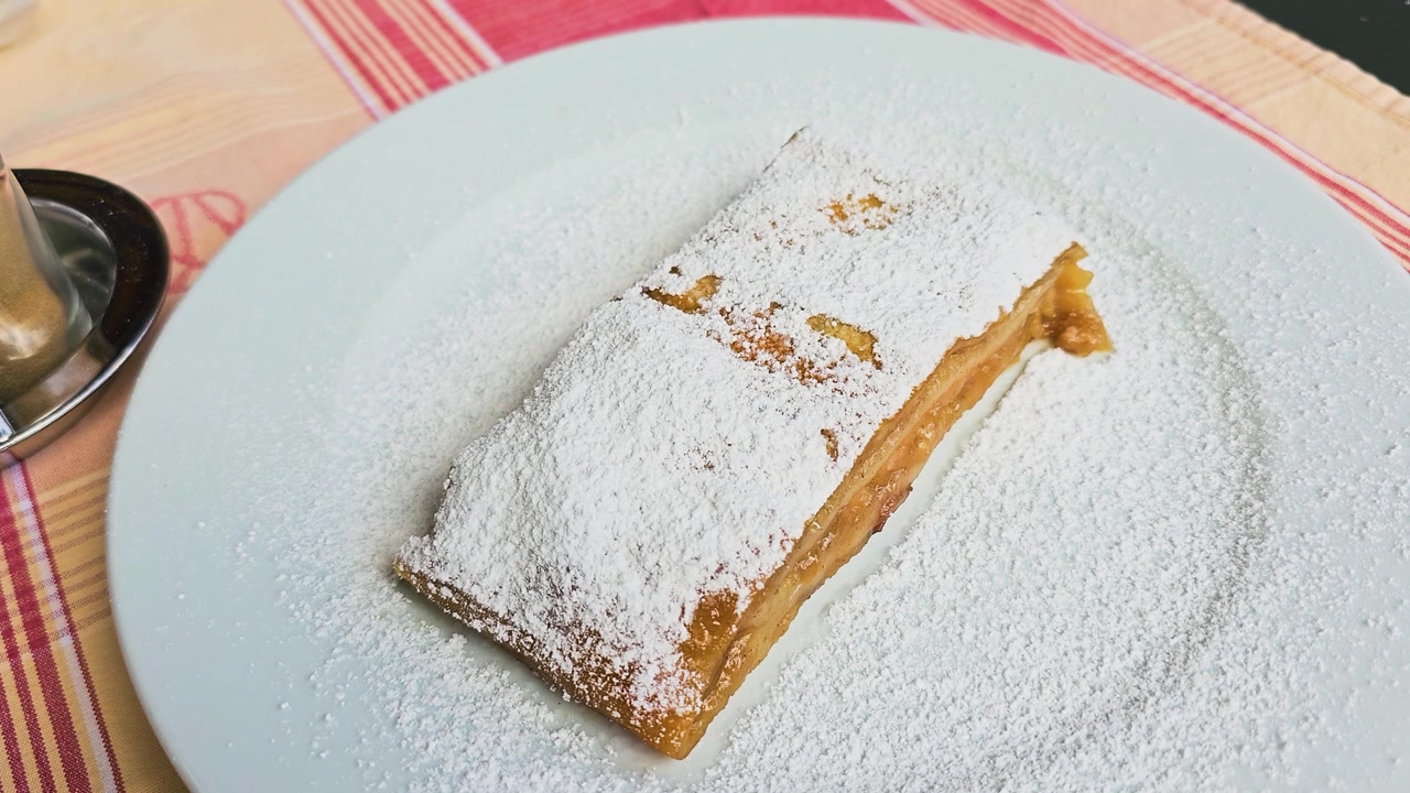 Güneşli bir Avrupa restoranının dış mekan teras masasında, beyaz bir tabakta pul pul dökülen altın rengi hamur işi, pudra şekeri ve tatlı elma dolgusu ile taze elmalı strudel tatlısı, klasik Avusturya kafe yemeği.