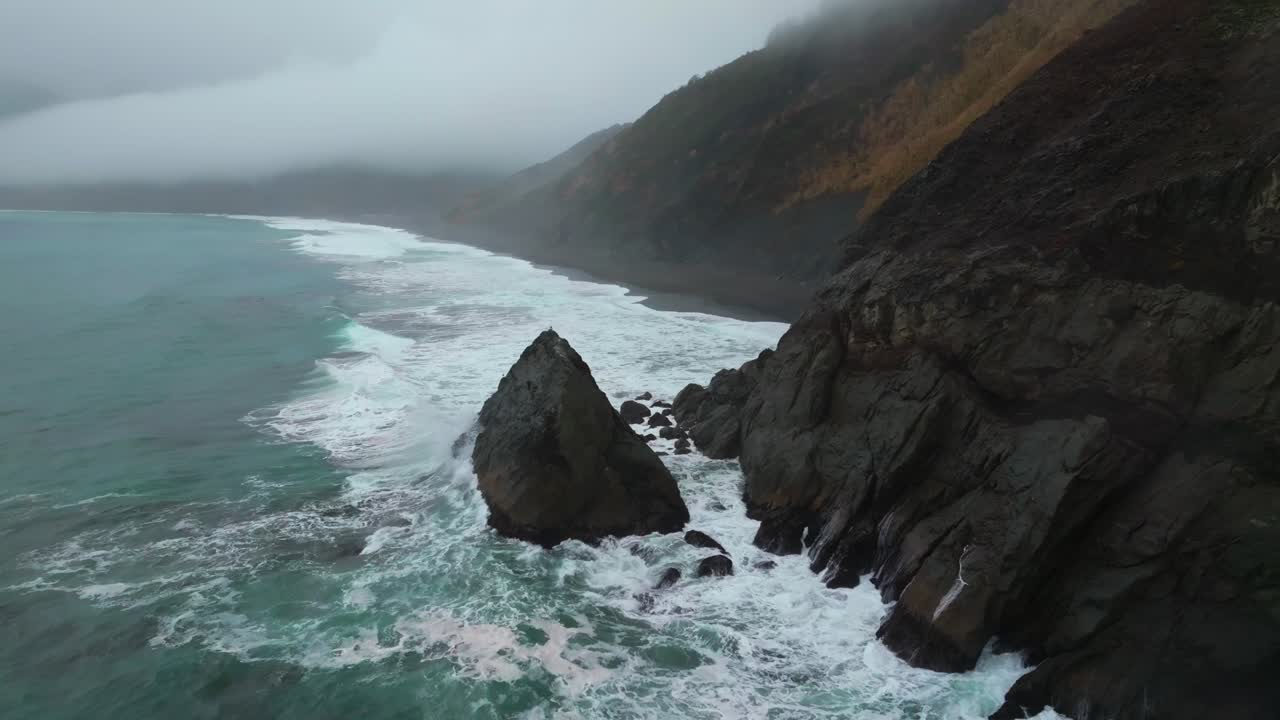 Rugged dramatic untouched coastline surf waves crashing boulder rocks USAL Beach campground cloud layer mist rain fog aerial drone California PNW Lost Coast Trail morning nature landscape backwards