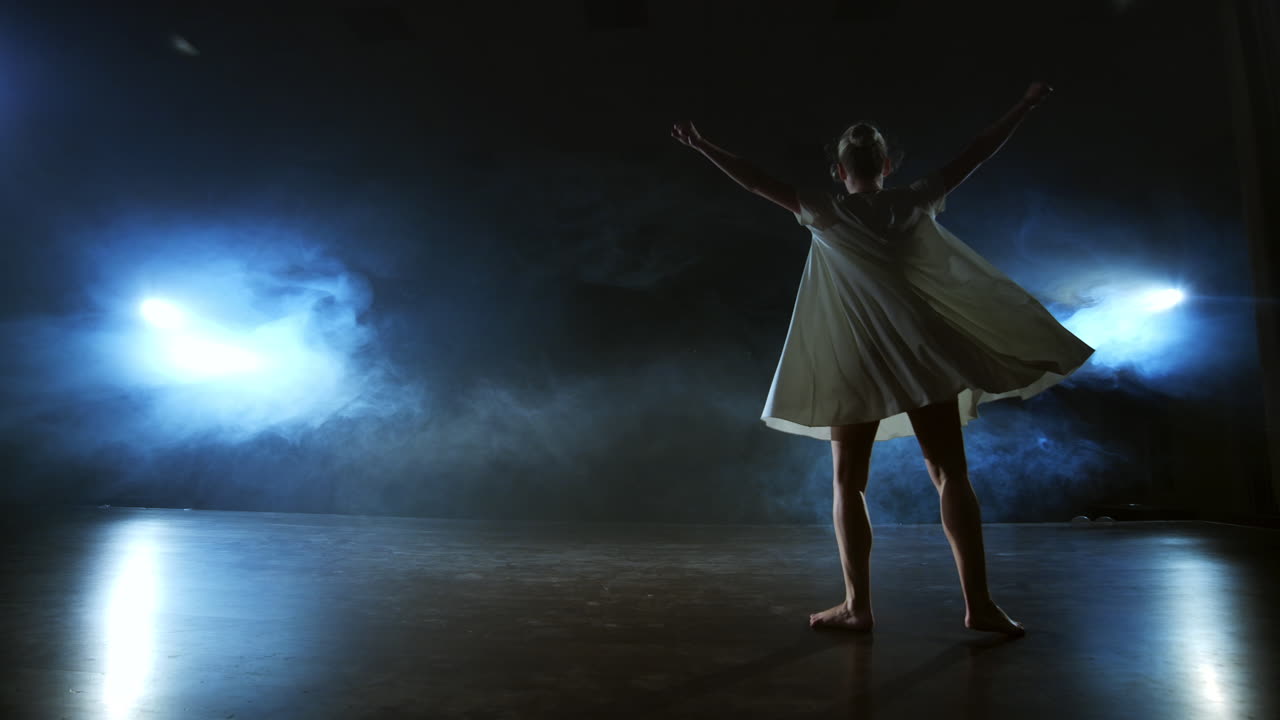 Modern dance girl in a white dress dances a modern ballet jumps on the stage with smoke in the blue spotlights.