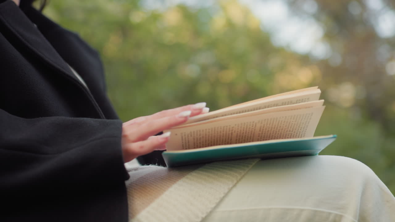 Lower view of stylish woman seated with legs crossed, reading through book on lap, hand steady on cover, pausing to pick up beverage beside bench for sip, calm autumn park greenery, soft daylight