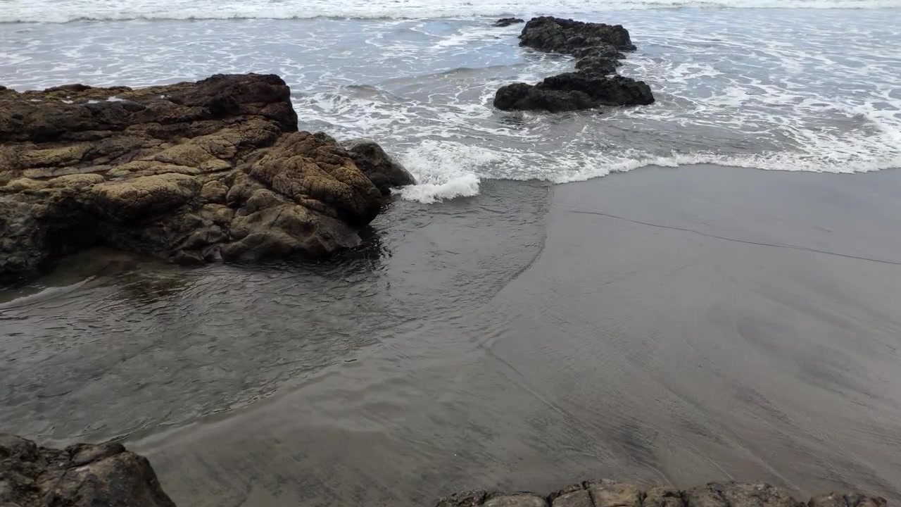 Onde che lambiscono delicatamente scogli costieri scuri lungo un litorale sabbioso.