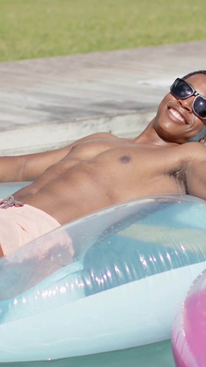Vertical video: Sun urging man in pink trunks sunglasses relaxing shifting on blue float pink ring