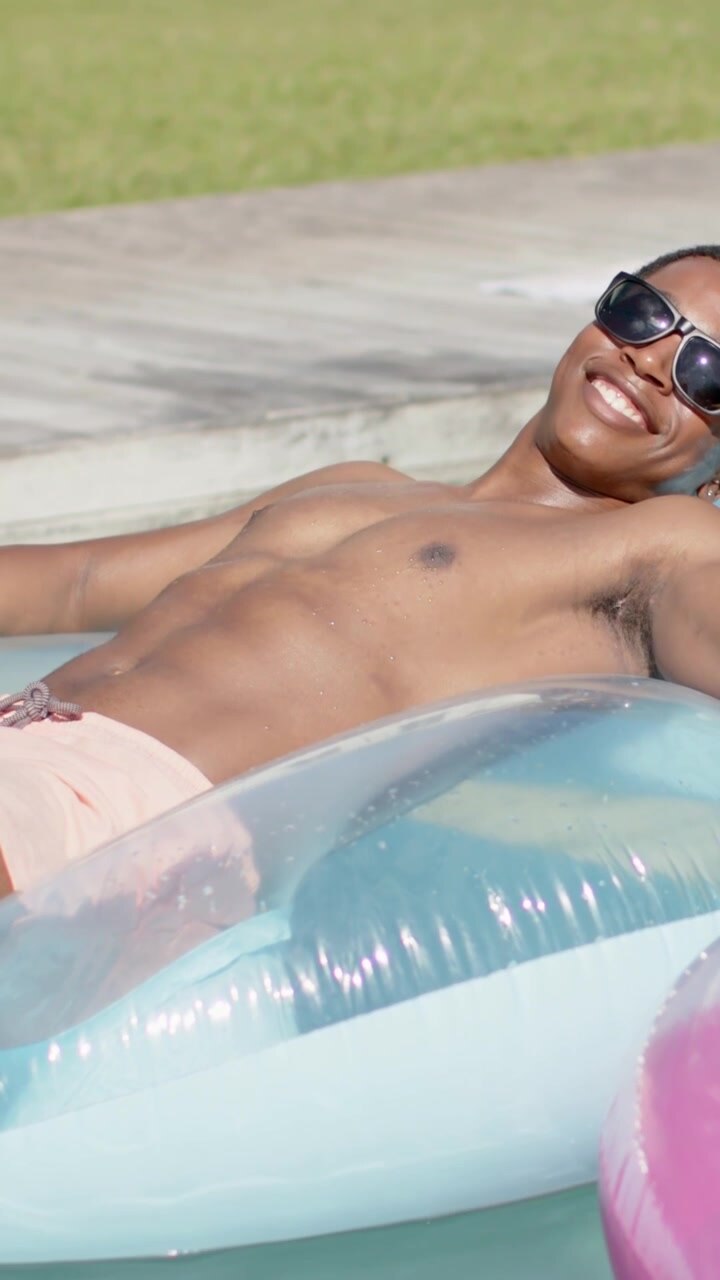 Vertical video: Sun urging man in pink trunks sunglasses relaxing shifting on blue float pink ring