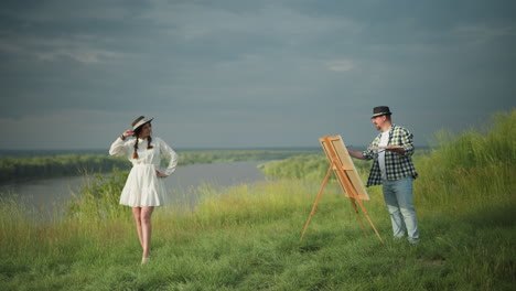 a woman in a white dress and hat stands poised in front of an artist wearing a checkered shirt, jeans, and a hat, who is holding a palette and painting her in a grassy field beside a lake