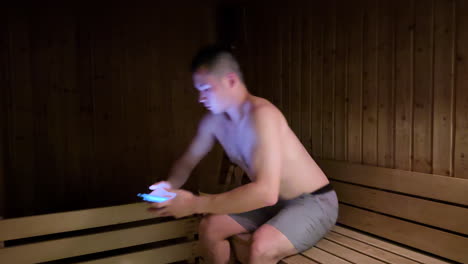 Asian-Millennial-Man-Entering-Wooden-Sauna-and-Sitting-Down-While-Using-Smartphone