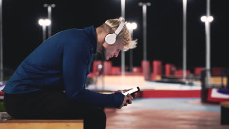 hombre rubio deportivo sentado en el parque escuchando música con auriculares bluetooth y usando el teléfono móvil mientras toma un descanso durante su sesión de entrenamiento en la noche 2