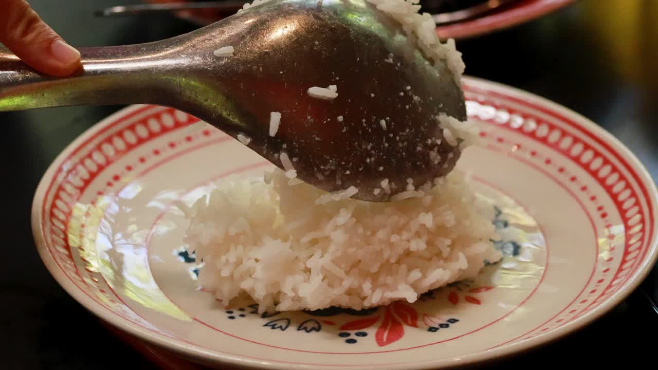 A spoon serves steamed rice onto a colorful plate, highlighting traditional Thai dining. Warm lighting enhances the inviting atmosphere