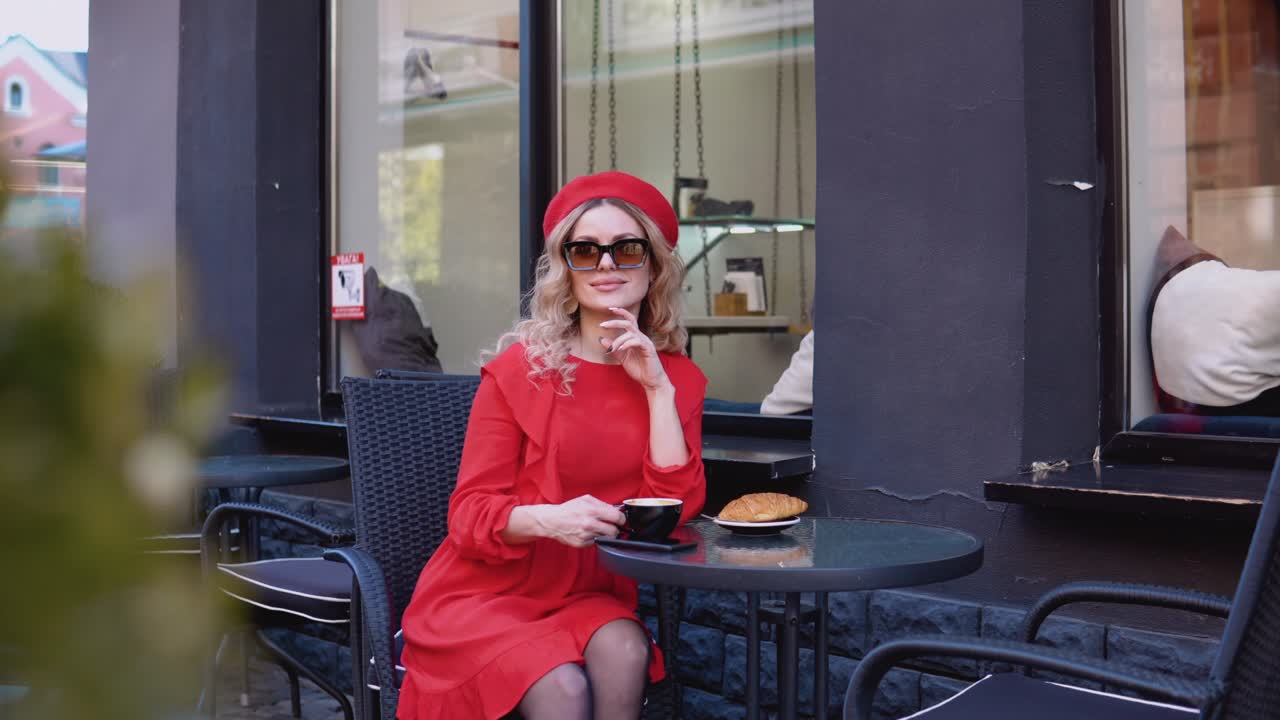 Slow morning aesthetic.Young romantic attractive woman smiling looking at camera. Woman in a red dress and beret sitting with a cup of coffee at a table on the street