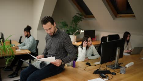 Focused caucasian young man sitting on the table with laptop and documents, solving the problem