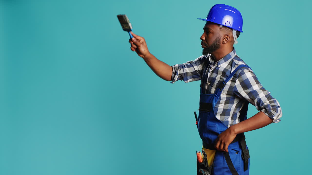 Young carpenter painting walls with paintbrush on camera