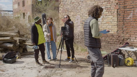 Tres-Compañeros-De-Producción-Hablando-Sobre-Un-Proyecto-De-Grabación-Y-Riéndose-Frente-A-Una-Cámara-En-Un-Trípode-En-La-Calle-Mientras-Otro-Trabajador-Toma-Notas-1