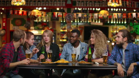 a multi-ethnic group of young men and women drink beer at a bar and eat chips and cheerfully debate about the university. laughing at a joke.