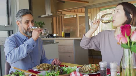 happy diverse couple sitting at table in dining room, eating dinner and drinking wine