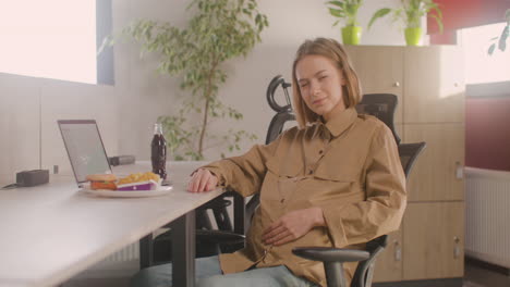 Arbeitende-Schwangere-Frau-Sitzt-Am-Schreibtisch-Und-Lächelt-In-Die-Kamera-Während-Der-Mittagspause-Im-Büro