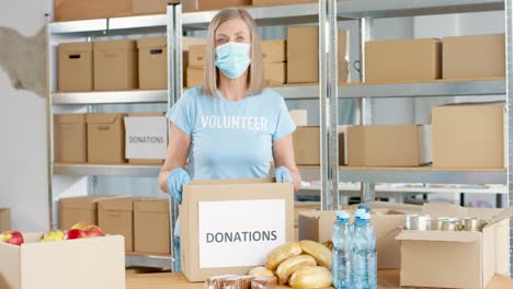 Mujer-Caucásica-Voluntaria-Con-Máscara-Facial-Empacando-Cajas-De-Donación-Y-Mirando-La-Cámara-En-Un-Almacén-De-Caridad