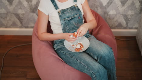 overhead view of person in blue denim overall seated on pink bean bag carefully peeling egg shell into white bowl placed on lap, with relaxed posture and scattered egg shells