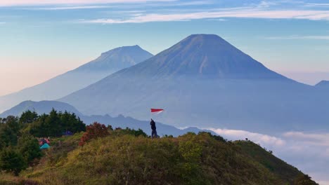 majestic mountain landscape with a person waving a red - white (indonesia) flag on a grassy hilltop above the clouds. the concept of pride in one's nation and state. aerial drone view
