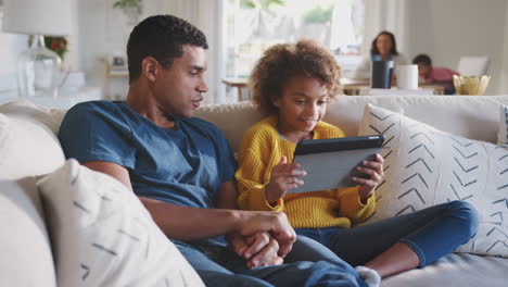 father and his pre-teen daughter on sofa using tablet computer, mum and toddler in the background