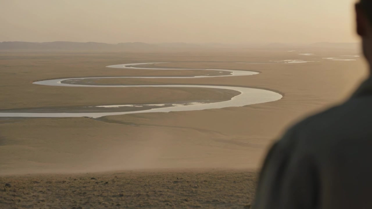 Camera shifting dark-coated man standing and watching winding river on dusty open plain, copy space