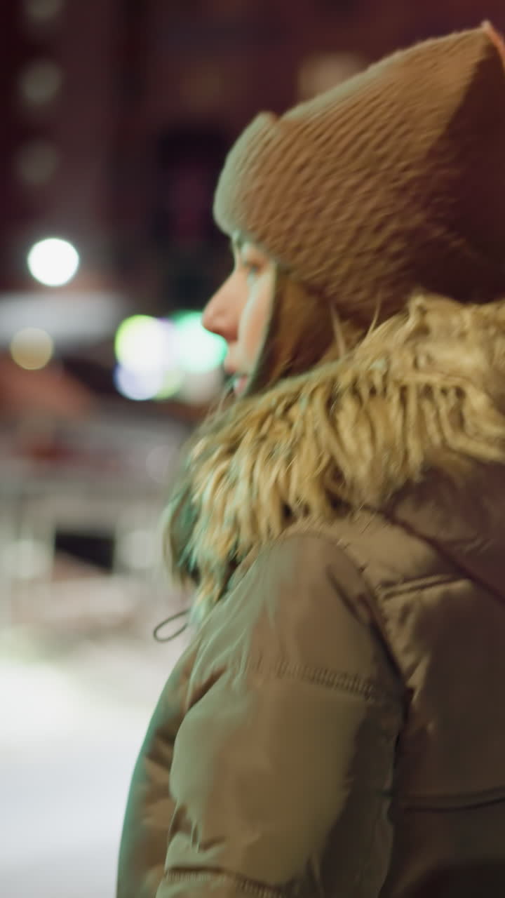 вид от первого лица женщины в зимнем пальто с меховым капюшоном и коричневым обогревателем головы, пересекающей дорогу ночью. в фоновом режиме видны размытые городские огни и машины