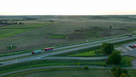 vista aérea de una carretera con un camión rojo viajando en ella, rodeado de vastos campos verdes y tierras de cultivo, durante la mañana temprano o tarde