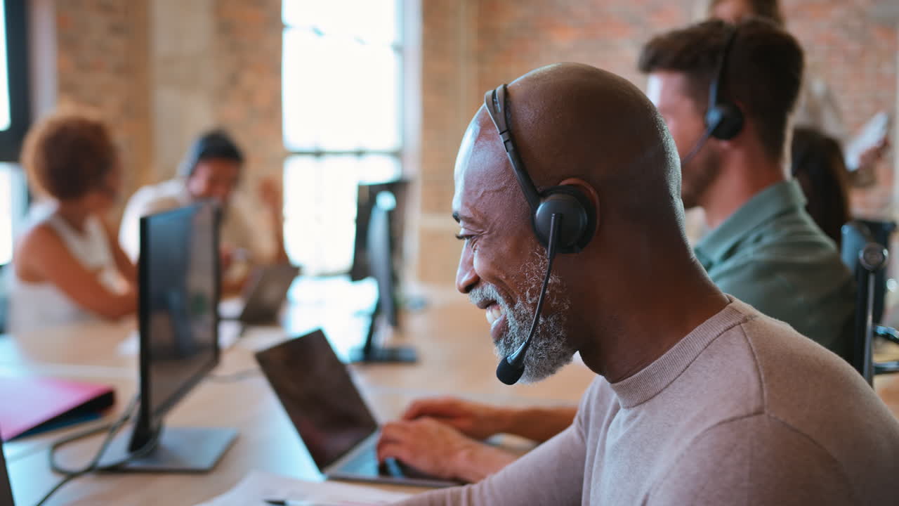 Portrait Of Mature Businessman In Multi-Cultural Team Wearing Headset In Customer Support Centre