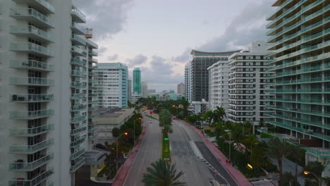 Multilane-trunk-road-lined-by-palm-trees-passing-through-modern-urban-borough.-Forwards-fly-along-multistorey-apartment-buildings-at-dusk.-Miami,-USA
