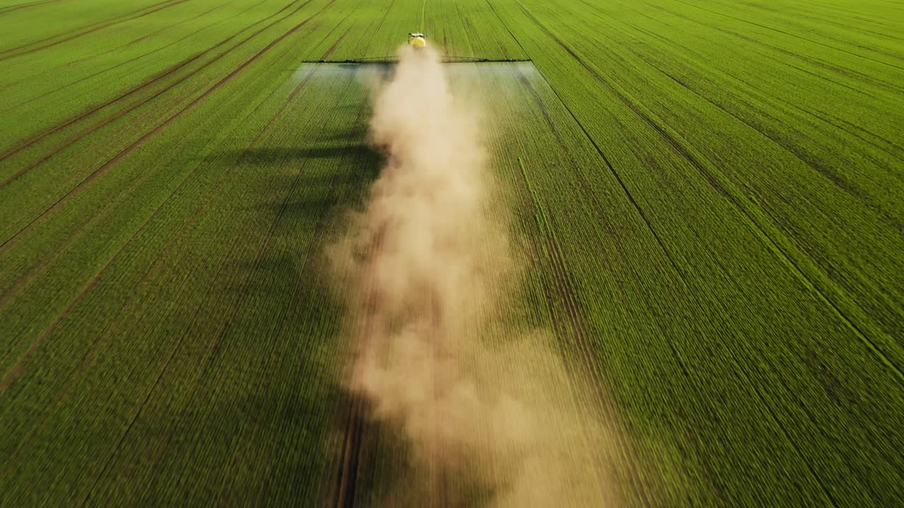 Aerial view of agricultural machinery spraying crops in a vast green field with dust cloud