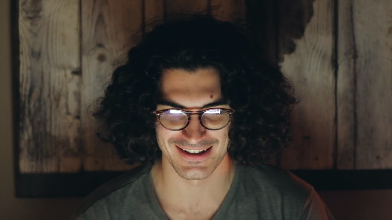 Joyful Curly-Haired Man Laughing at Social Media Content, Glowing Screen Light