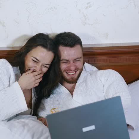 Joyful young couple on bed. Handsome man and attractive woman in white bathrobes laying on bed and laughing while looking into laptop. Happy together