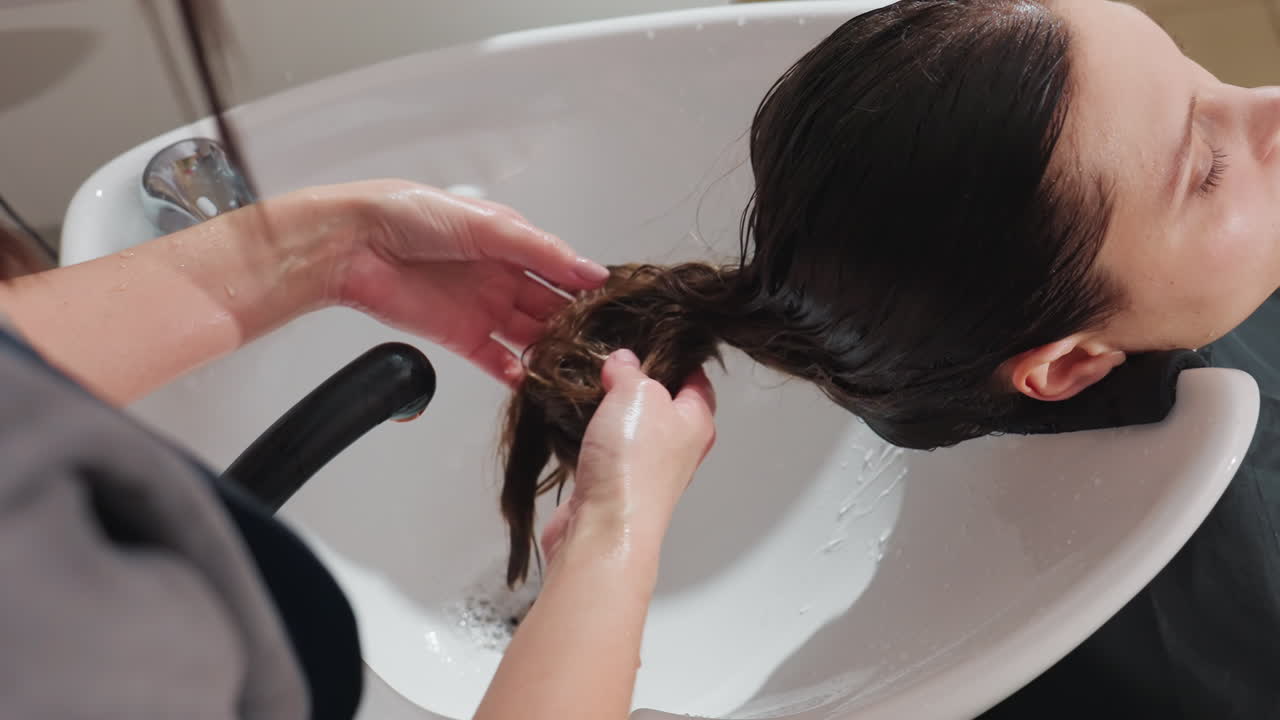 Donna sdraiata al lavandino del salone, parrucchiere che massaggia i capelli nel lavabo mentre applica shampoo e balsamo, movimenti lenti e rilassanti, capelli bagnati e scuri che gocciolano nella ciotola bianca, atmosfera rilassante