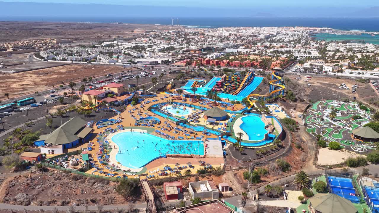 Acua Water Park in Corralejo with winding slides, pools, mini-golf, and palm-lined paths set against arid terrain showing organized recreation in northern Fuerteventura, Drone establishing shot