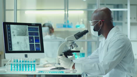masked lab scientist working with microscope and computer