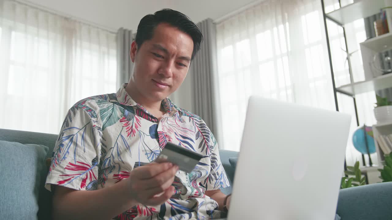 Asian Man Sitting On Couch In Living Room Holding Credit Card And Using Laptop Computer Shopping Online