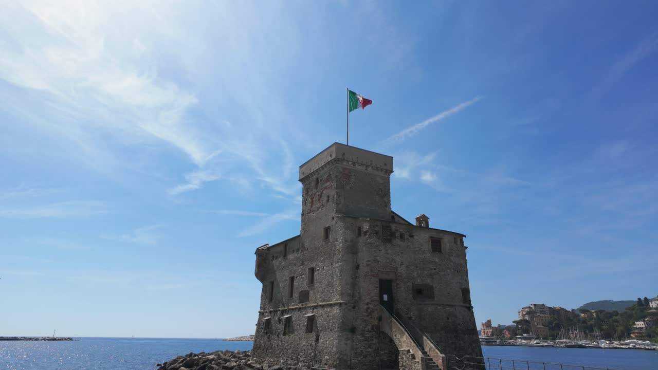 Video drone cattura lo storico Castello di Rapallo sulla costa ligure, bandiera italiana che sventola in cima alla torre di pietra, cielo azzurro vivido, mare calmo e costa illuminata dal sole, nessuna persona visibile, ripresa grandangolare