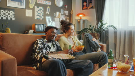 young couple enjoying snacks and drinks, watching tv and laughing at home