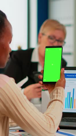 vertical video black businesswoman sitting at conference desk holding smartphone with green screen