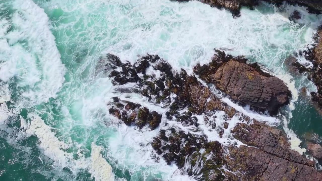 Ripresa cinematografica dall'alto con drone di potenti onde marine turchesi che si infrangono su scure rocce costiere coperte di alghe marine, sulla costa selvaggia e incontaminata dell'isola di Chiloé nella Patagonia cilena