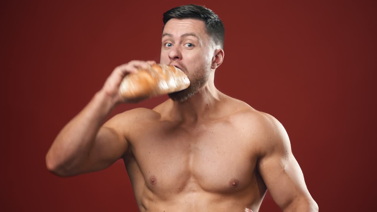 Hungry strong male eating unhealthy food. Half-length portrait of muscular man without shirt eats bread and sausage quickly on the studio background.
