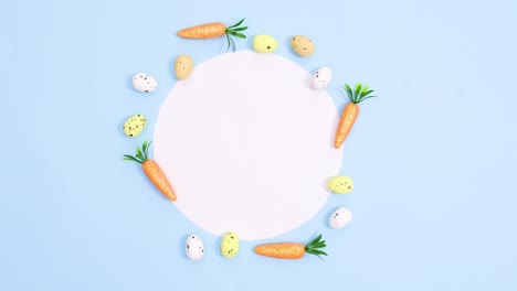 easter eggs and carrots appear around paper card note on pastel bright blue background. minimal flat lay stop motion