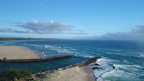 Slow-aerial-view-of-a-beach-at-sunrise-with-nice-rolling-waves-in-the-blue-ocean,-white-sand-beach-and-rock-jetty