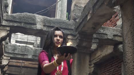 beautiful-Indian-girl-in-old-stepwell-wearing-traditional-Indian-red-saree,-gold-jewellery-and-bangles-holding-fire-plate